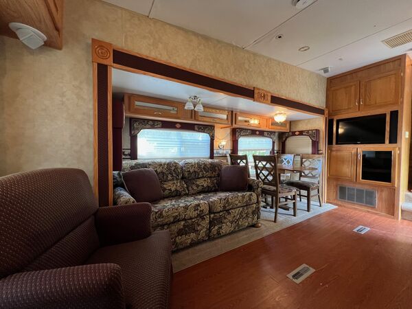 Living room with a recliner, a patterned sofa in the slide-out, a dining table with chairs, and a television built into the oak cabinetry