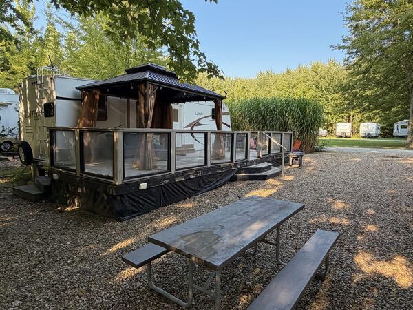 Long glass-railed deck with a curtained gazebo along the side of the trailer, and a picnic table on the gravel in front