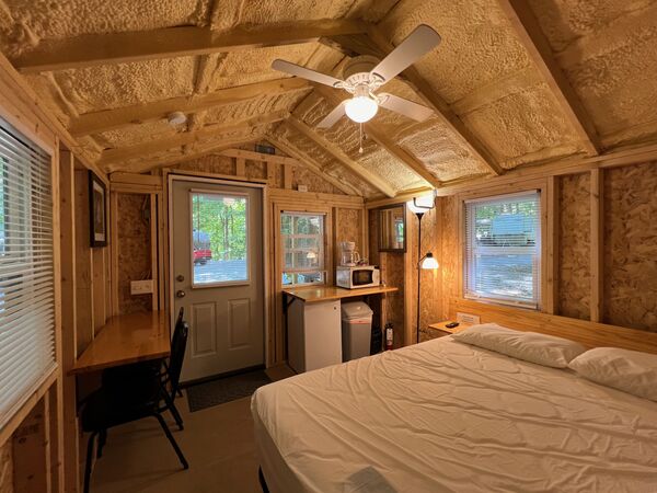 Interior with a queen bed, a long wooden counter with chairs under the window, a kitchenette with a microwave and coffee maker, and a ceiling fan