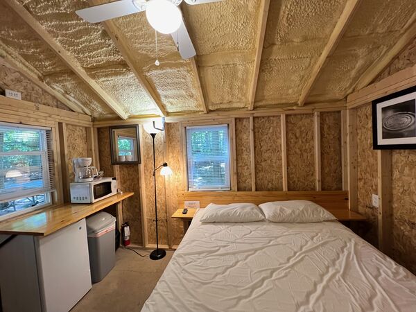 Queen bed against a wooden headboard under a window, with a floor lamp, a mirror, and a counter with a microwave and coffee maker above a mini fridge