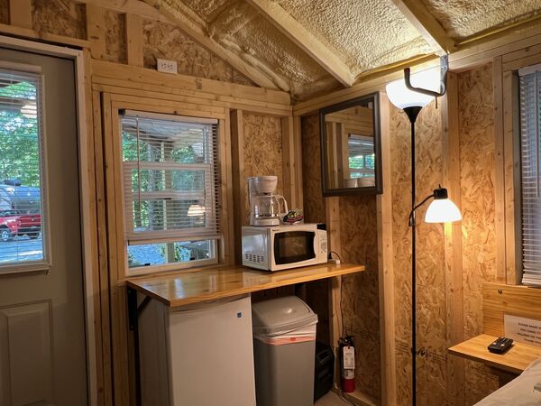 Kitchenette counter under a window with a coffee maker and microwave above a mini fridge and trash can, a mirror and floor lamp beside it