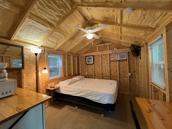 Queen bed beneath a ceiling fan, with a wall air conditioner, mounted television, floor lamp and windows on both sides