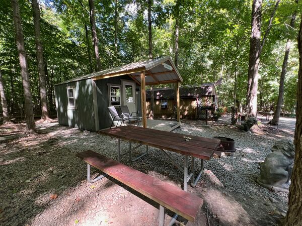 Side view of the green cabin and its covered porch with two chairs, a red picnic table in front and a stone lion statue at the edge of the wooded site