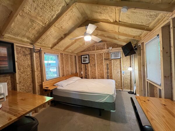 Queen bed under a ceiling fan with a wall air conditioner and mounted television, long wooden counters along both walls