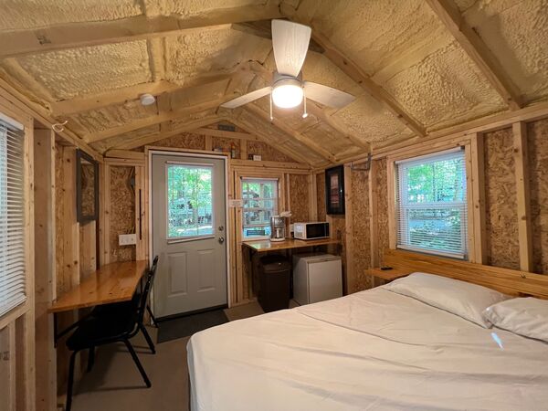 From the bed toward the front door: a long counter with a chair, a kitchenette with a coffee maker, microwave and mini fridge, and a ceiling fan