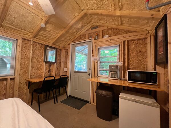 Kitchenette counter under a window with a coffee maker and microwave above a mini fridge, beside the front door and a counter with two chairs