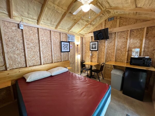 Queen bed with a red cover and two pillows, a small table with two chairs, a mounted television, and a counter with a microwave and coffee maker above a mini fridge